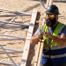 A construction worker with a beard, grey helmet, cool sunglasses and a neon vest standing in dirt while holding a cable